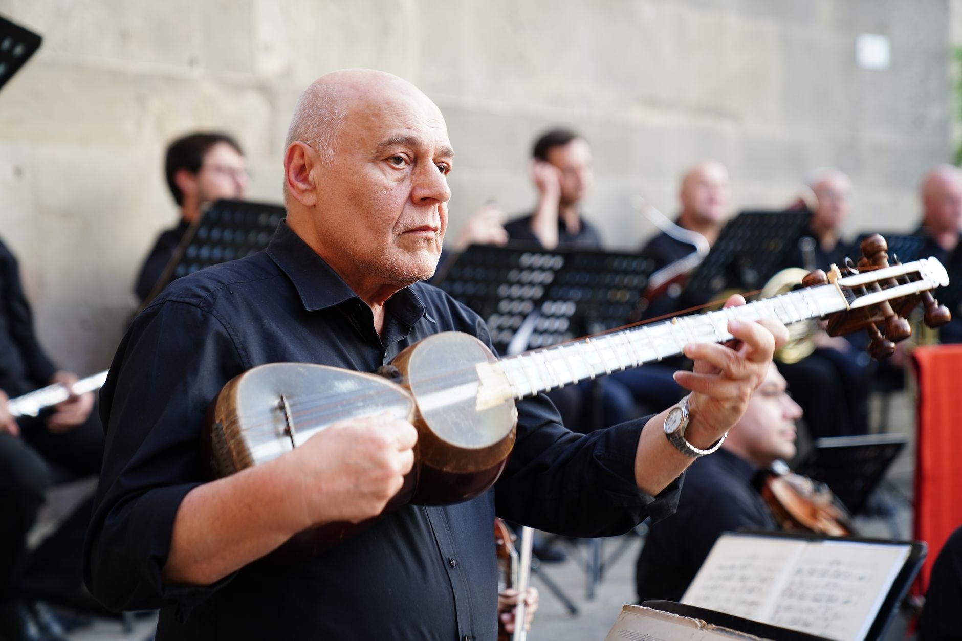 Elderly musician passionately playing a tar in an outdoor concert with an orchestra.