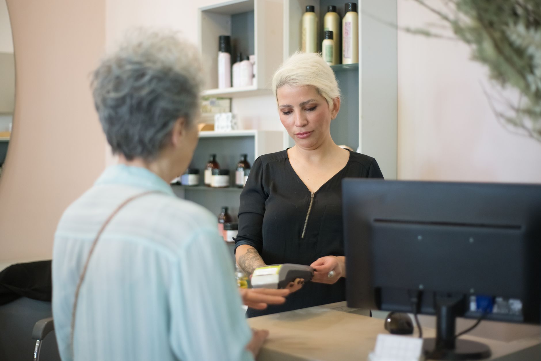 Two women at a hair salon checkout, one making a payment using a card reader.