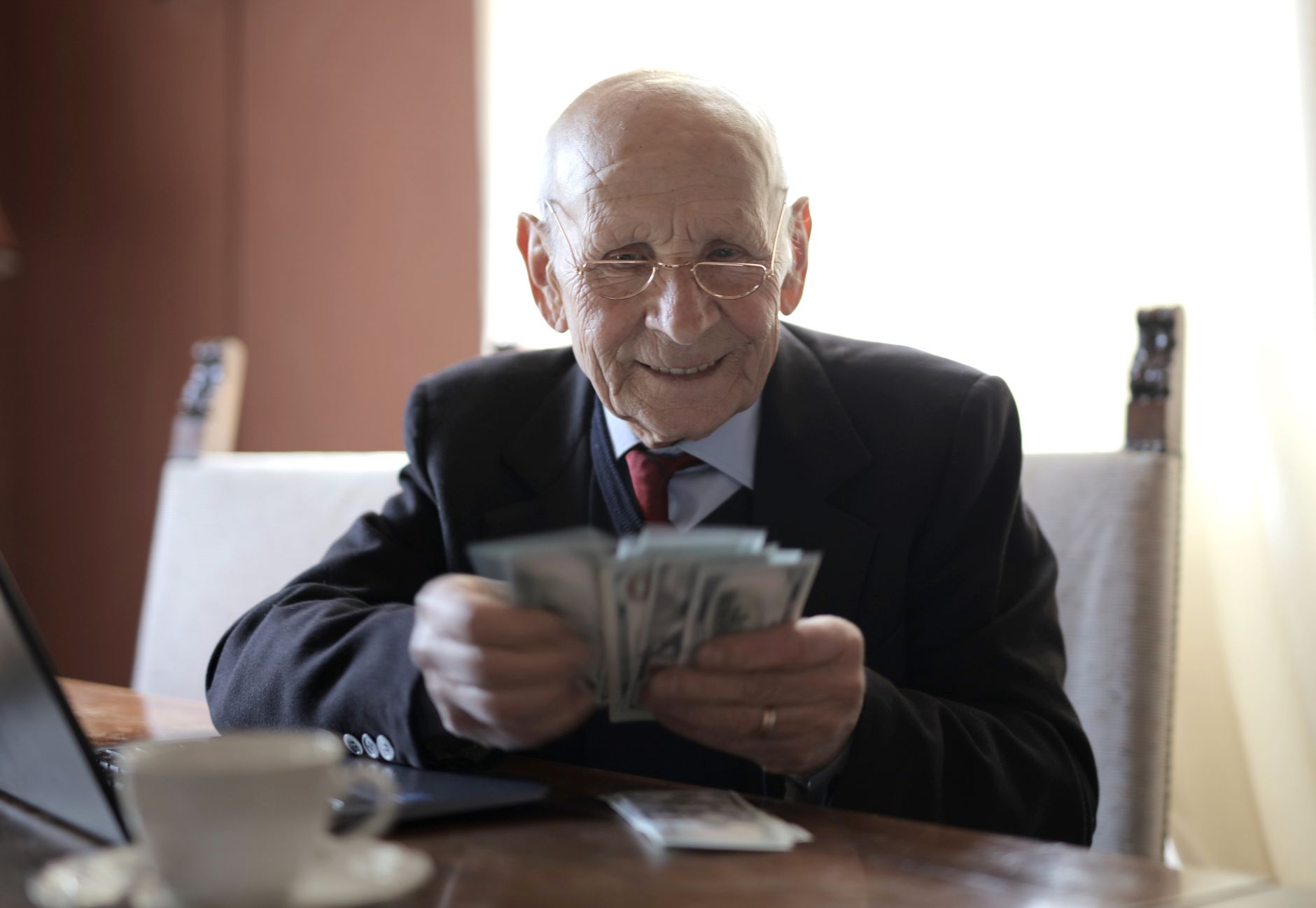 Positive senior businessman in formal suit and eyeglasses counting money bills while sitting at wooden table with cup of beverage and near o
