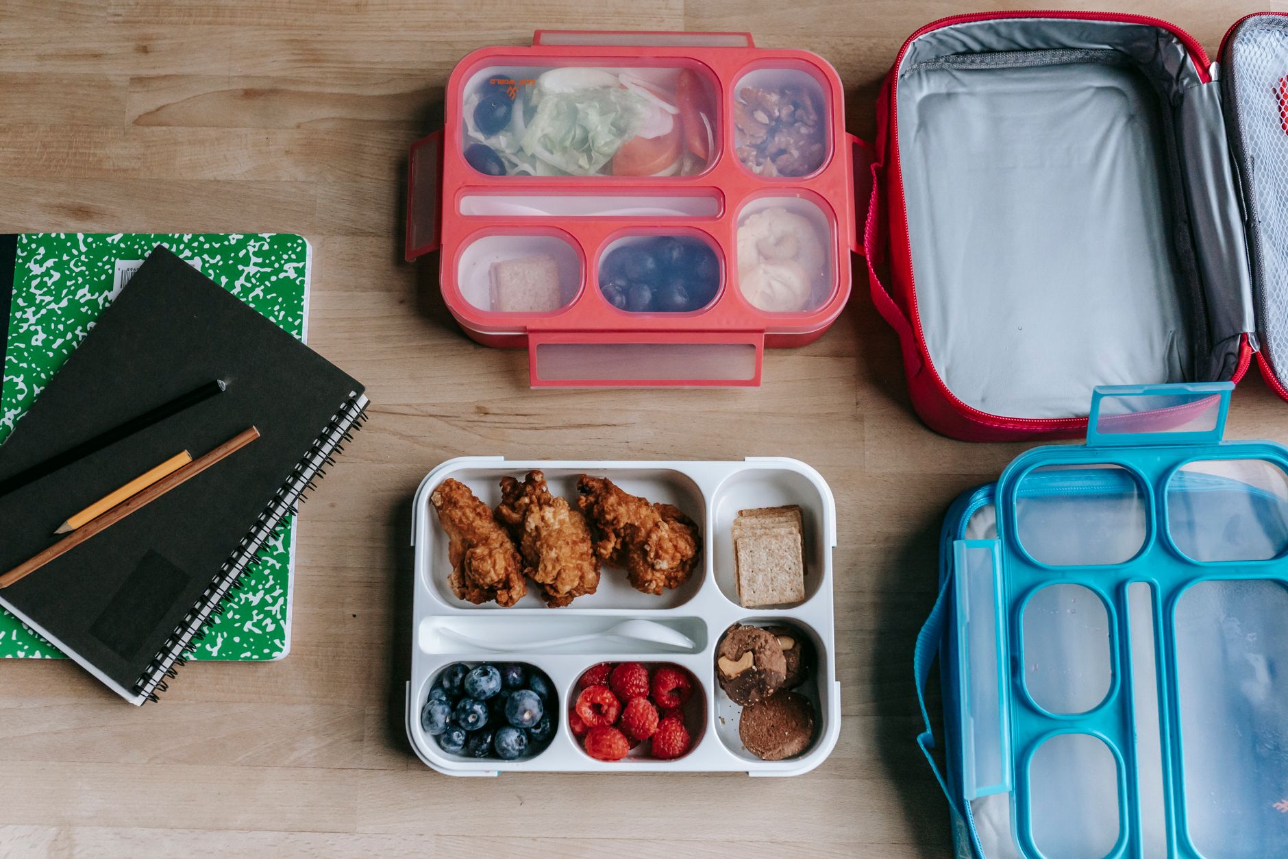 Top view of composed plastic lunchboxes near copybooks under pencils on wooden table