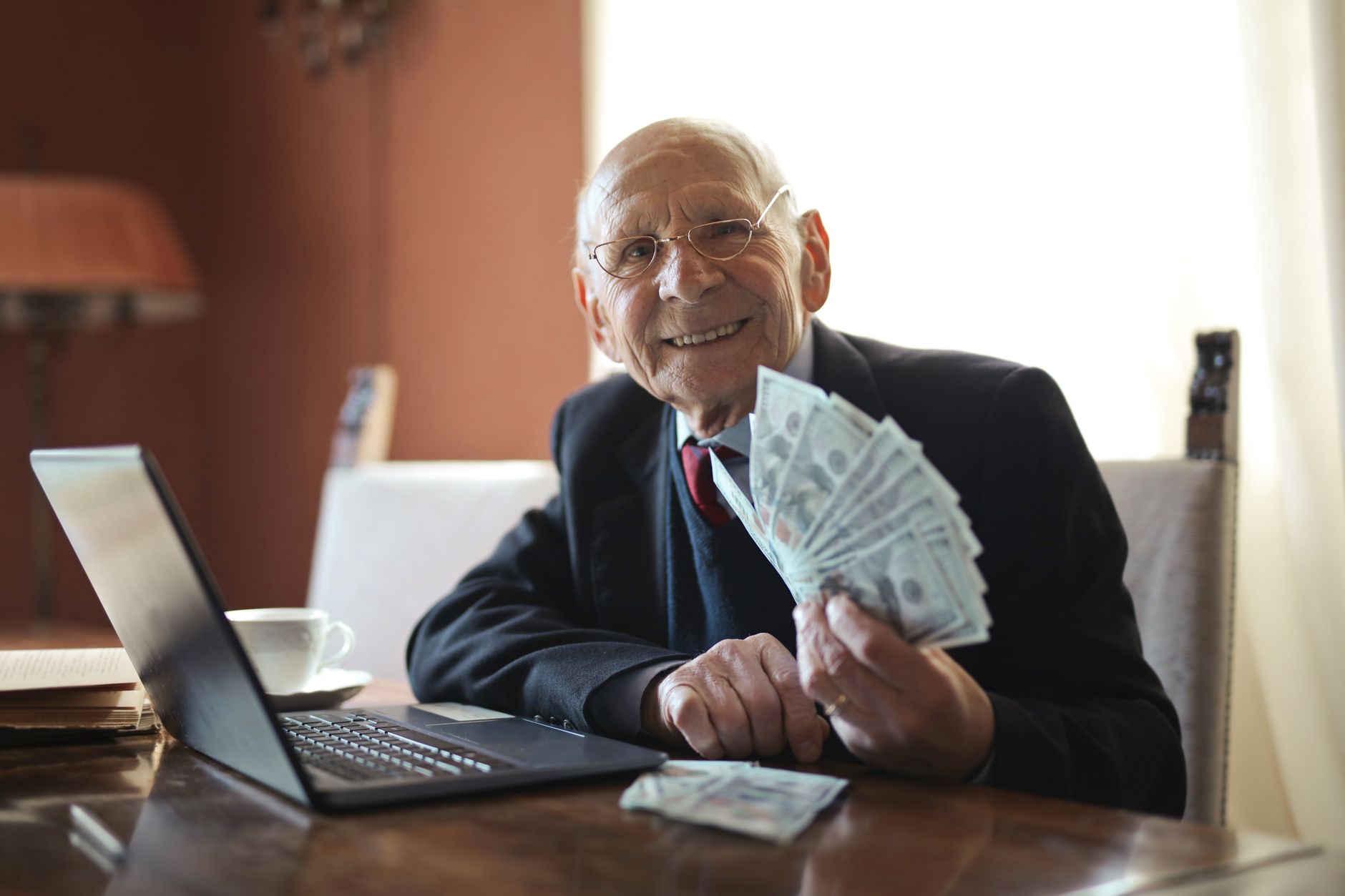 Positive senior businessman in formal suit and eyeglasses holding bunch of bills while working on laptop at table and looking at camera