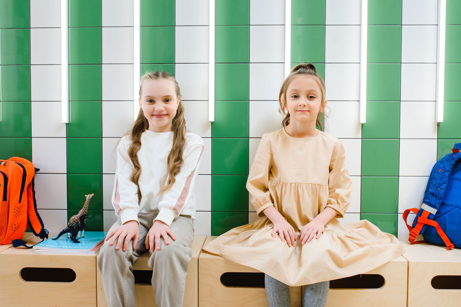 Two young girls sitting indoors in a school setting with colorful backpacks and a giraffe toy.