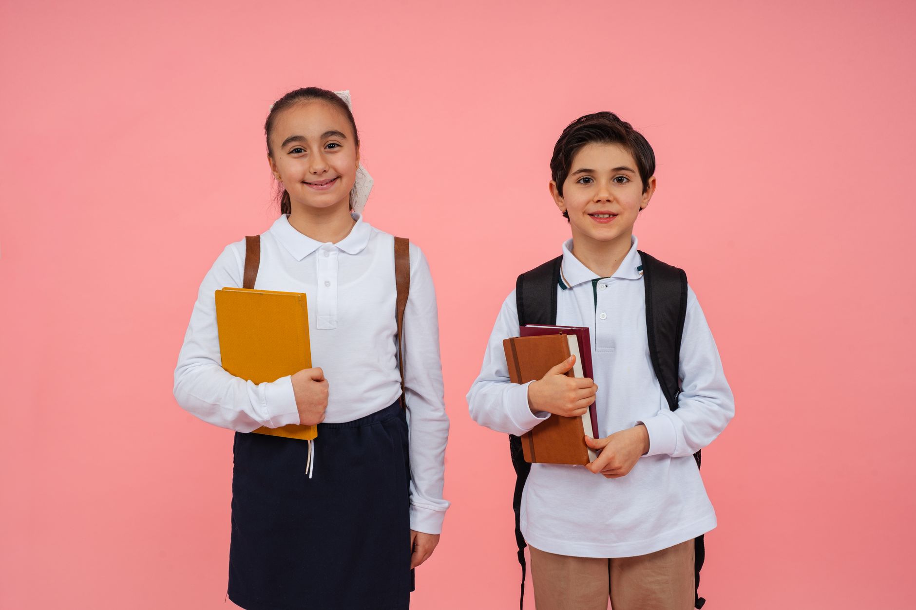 Two smiling school children holding books against a pink background, ready for school.