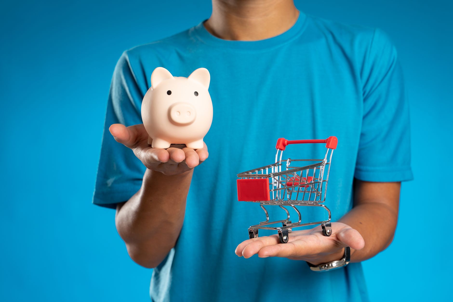 Man in blue shirt holding piggy bank and mini shopping cart, symbolizing savings versus spending.