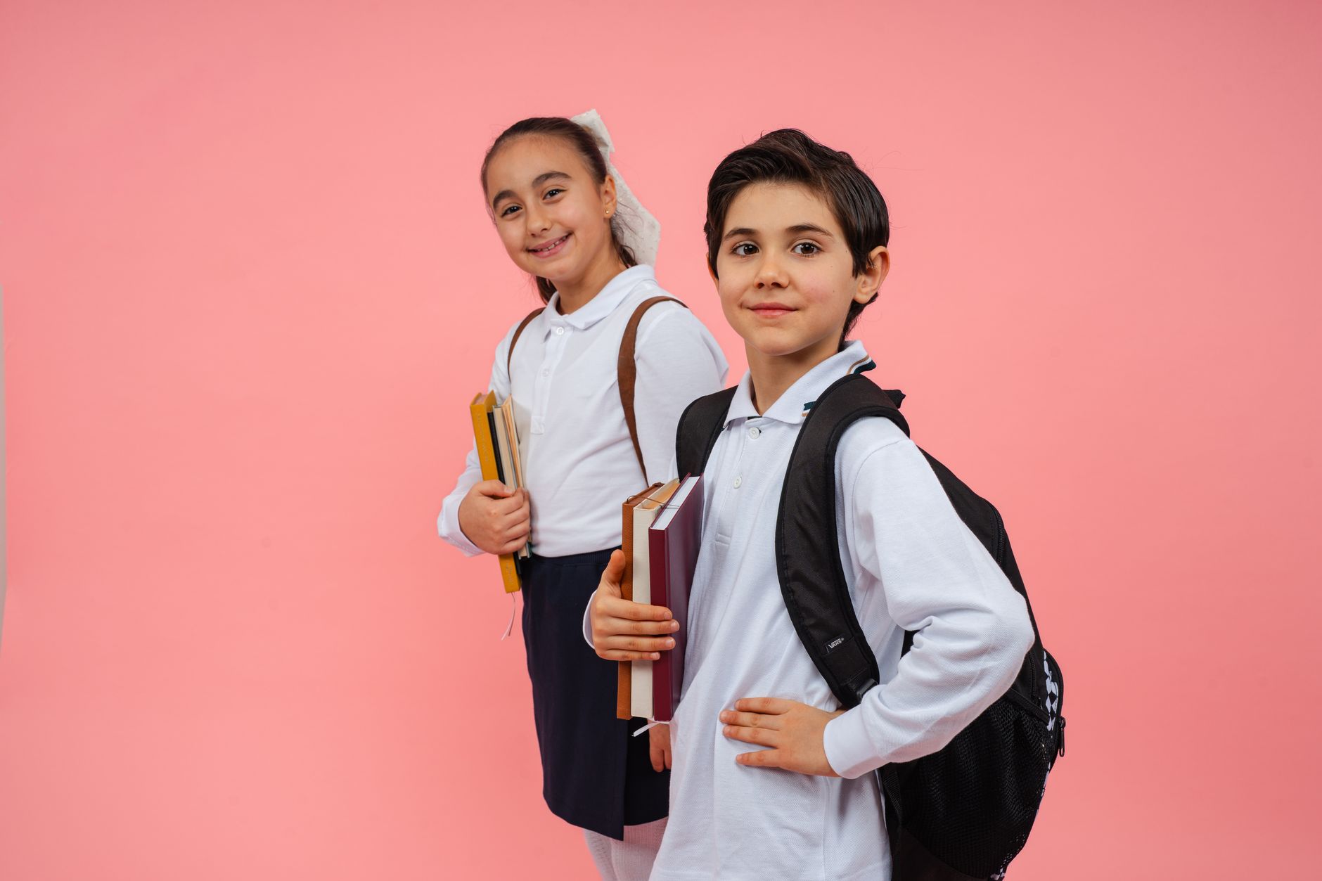 Two children in school uniforms holding books and backpacks against a pink backdrop.