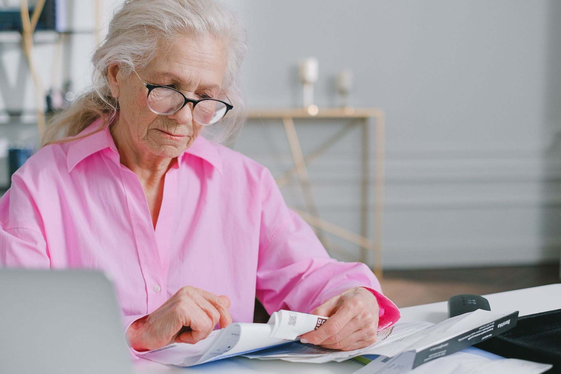 Elderly woman in pink shirt reading documents at a desk indoors.
