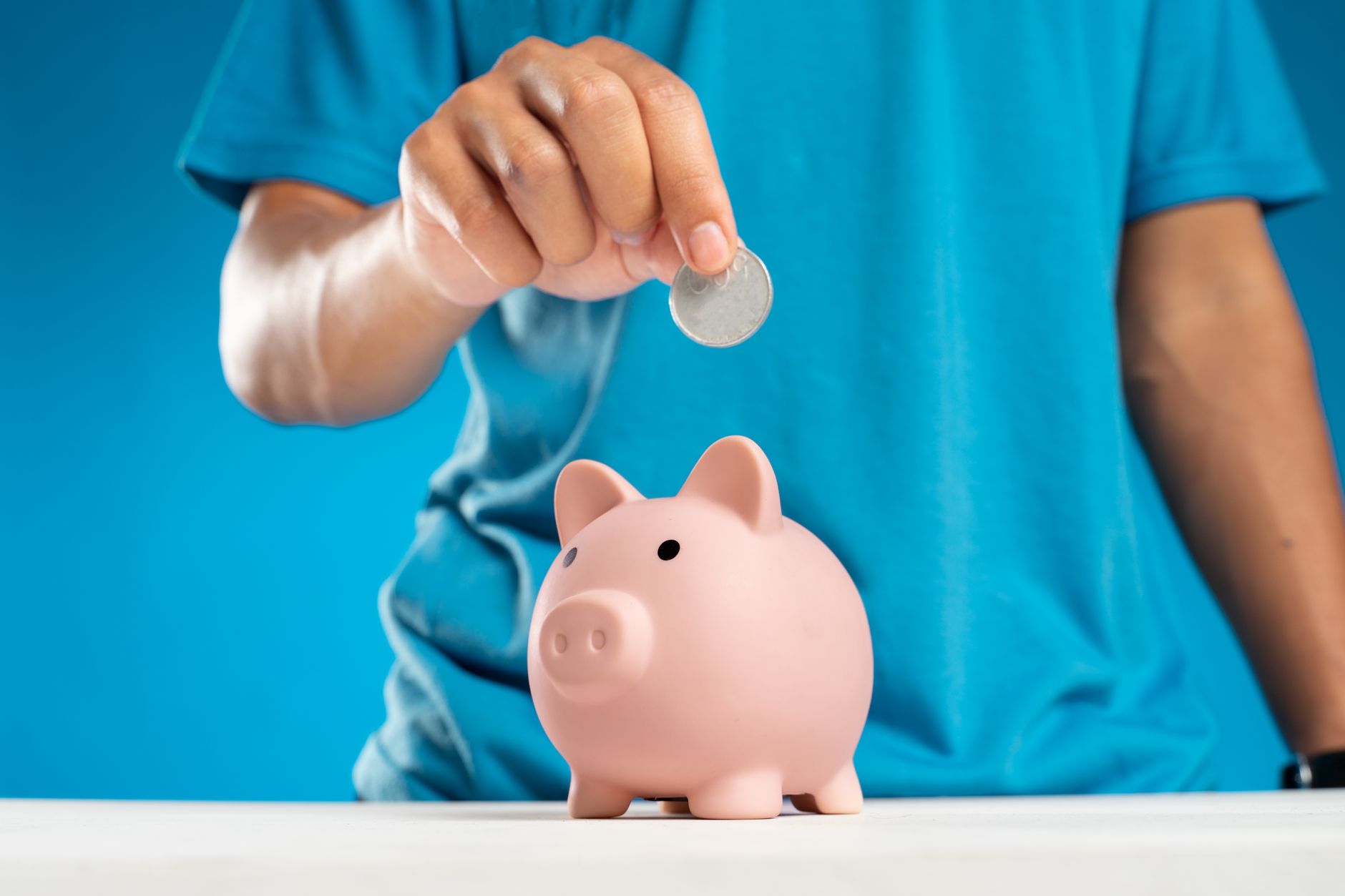 A close-up of an adult's hand dropping a coin into a piggy bank, symbolizing savings and investment.
