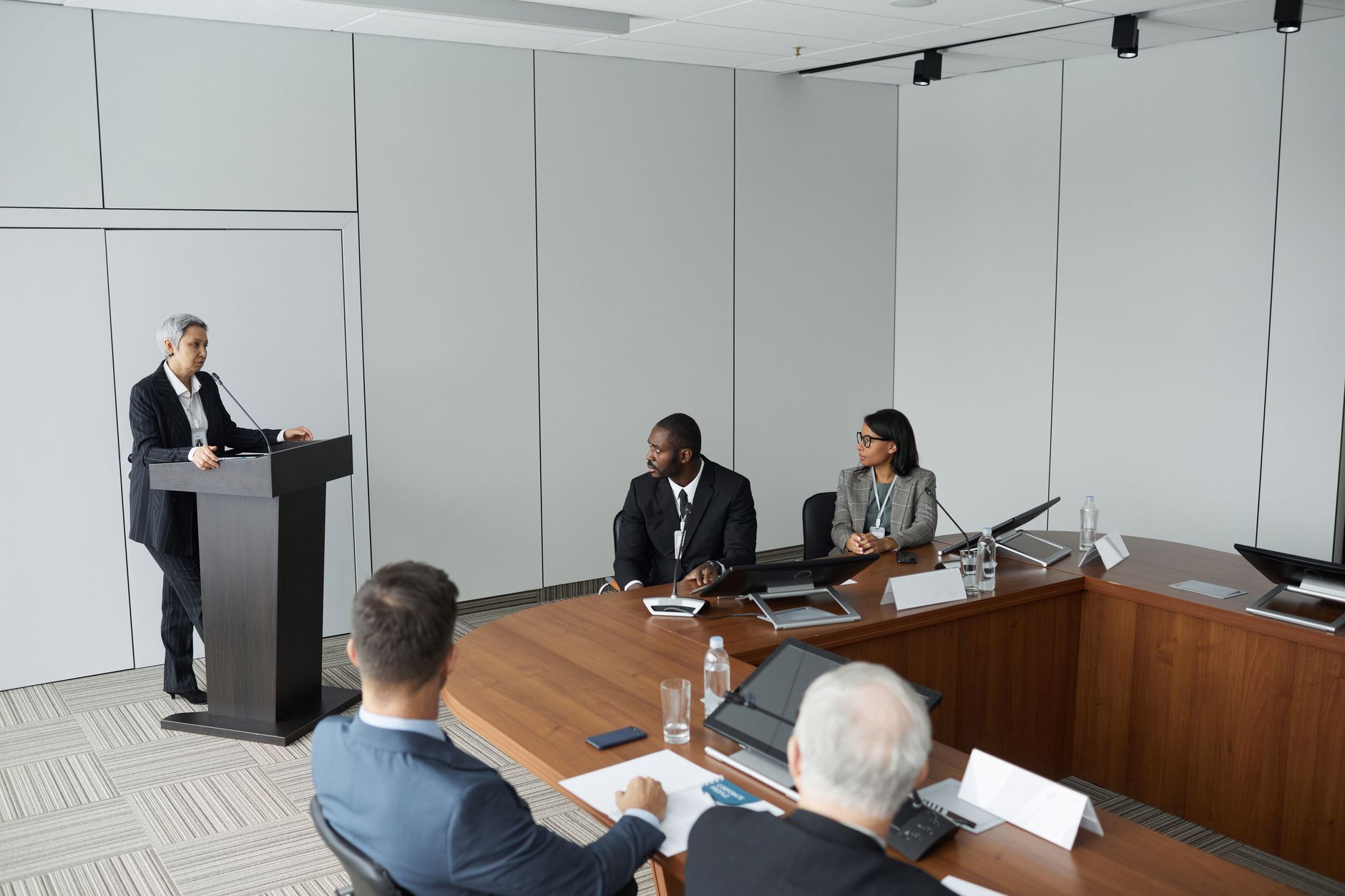 Diverse group of professionals in a formal meeting setting with a speaker at the podium.