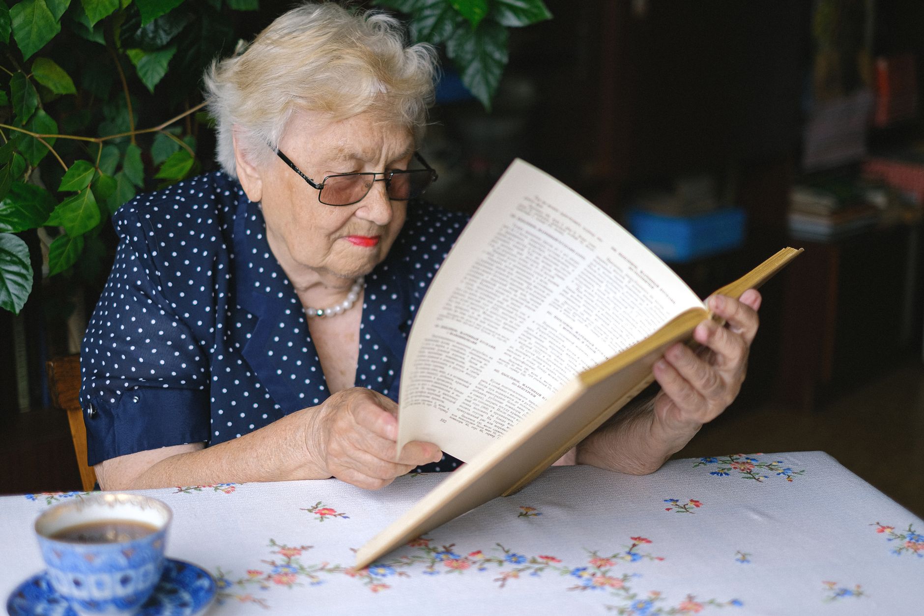 Serious female pensioner in eyeglasses reading book while sitting at table with cup of hot drink in room on blurred background