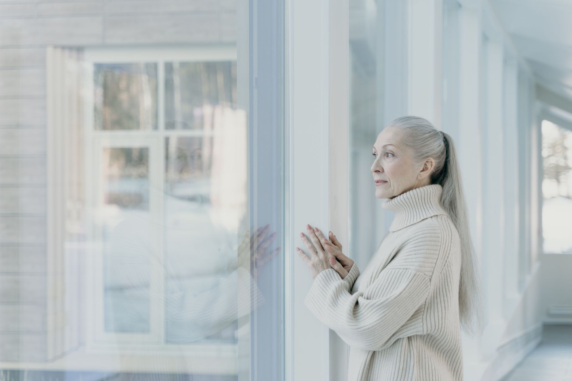 Serene elderly woman with grey hair looking out the window in a bright room, wearing a cozy sweater.