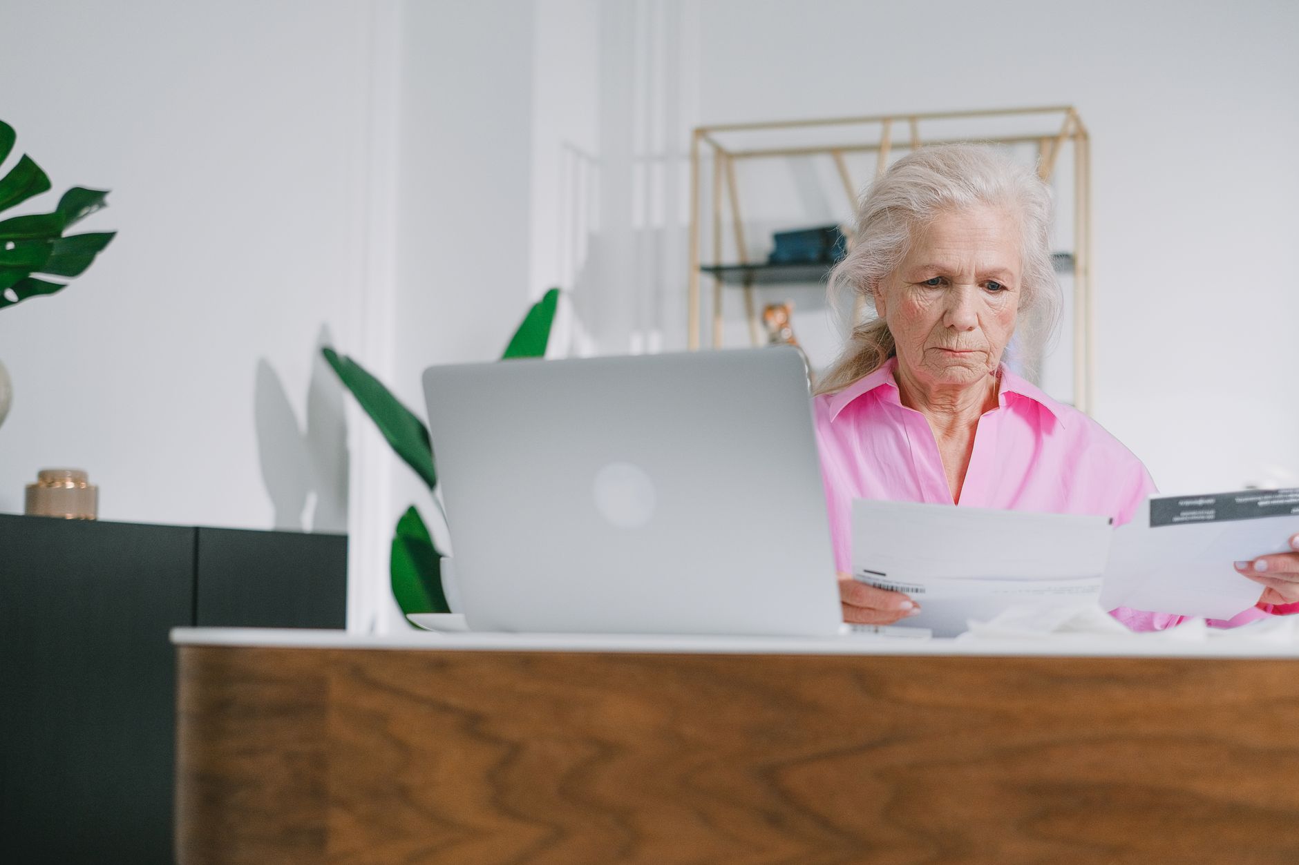Elderly woman focused on paperwork at home office with laptop.