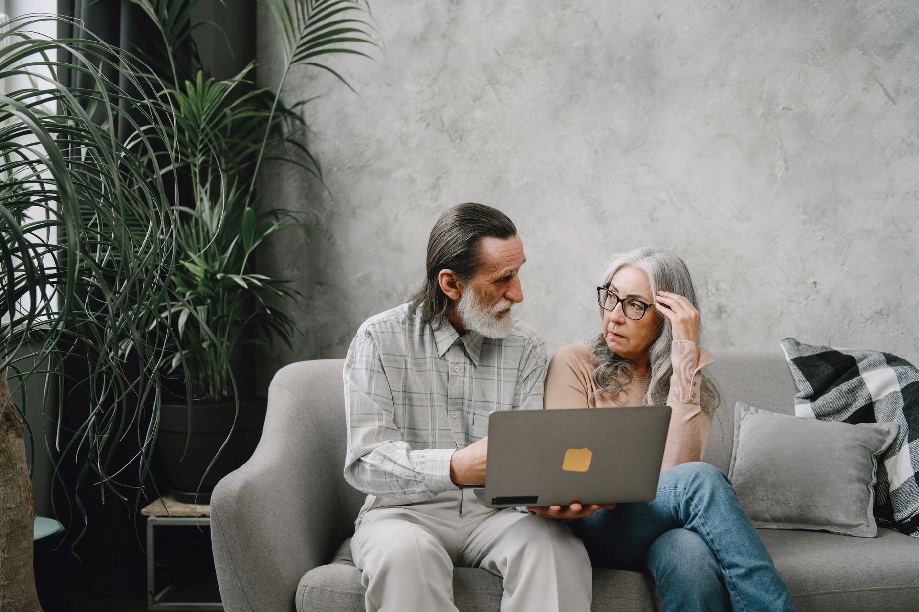 Elderly couple discussing while using laptop at home on sofa, surrounded by plants.