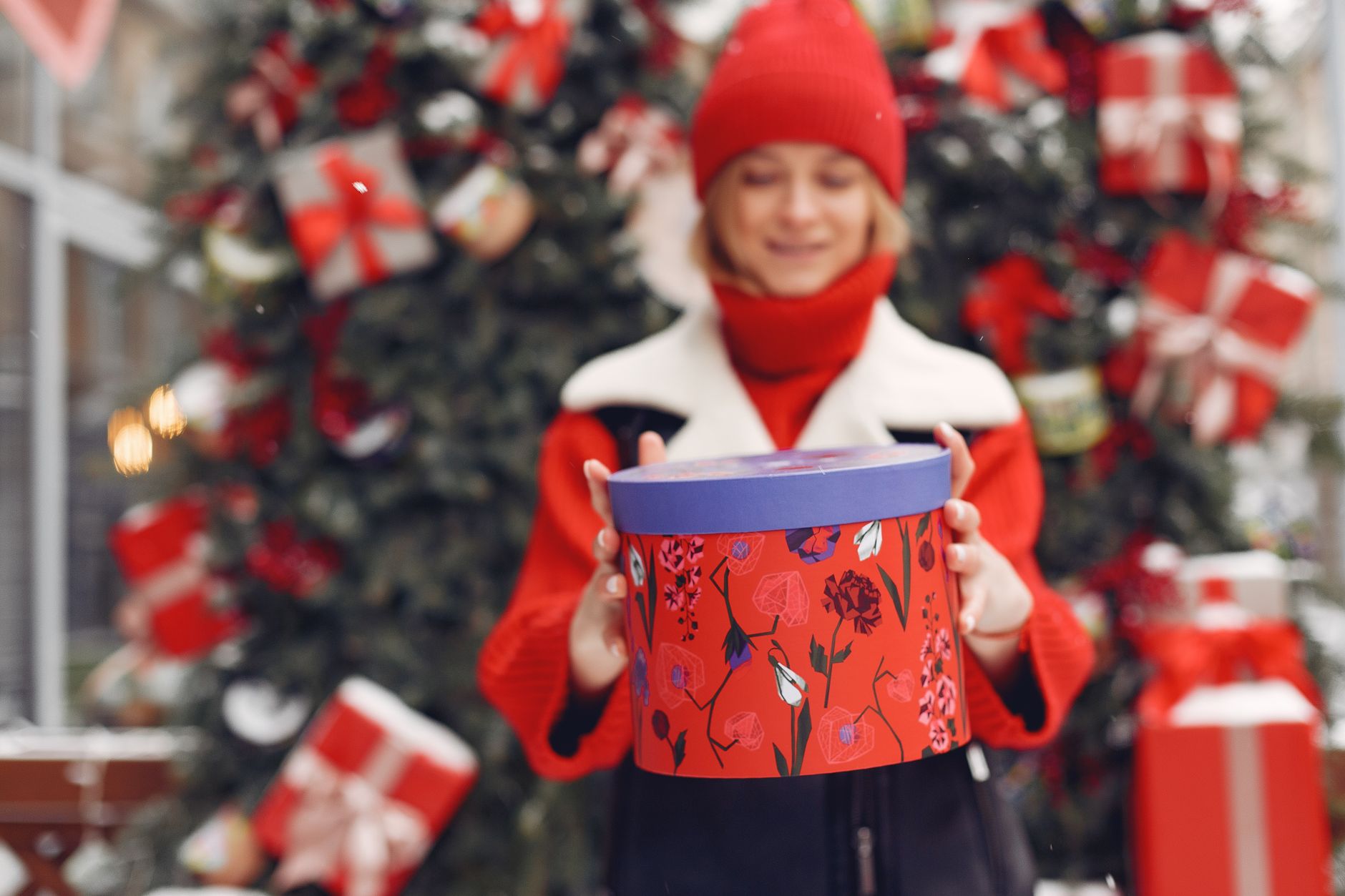 Woman in winter attire holding a vibrant gift box with Christmas tree in background.