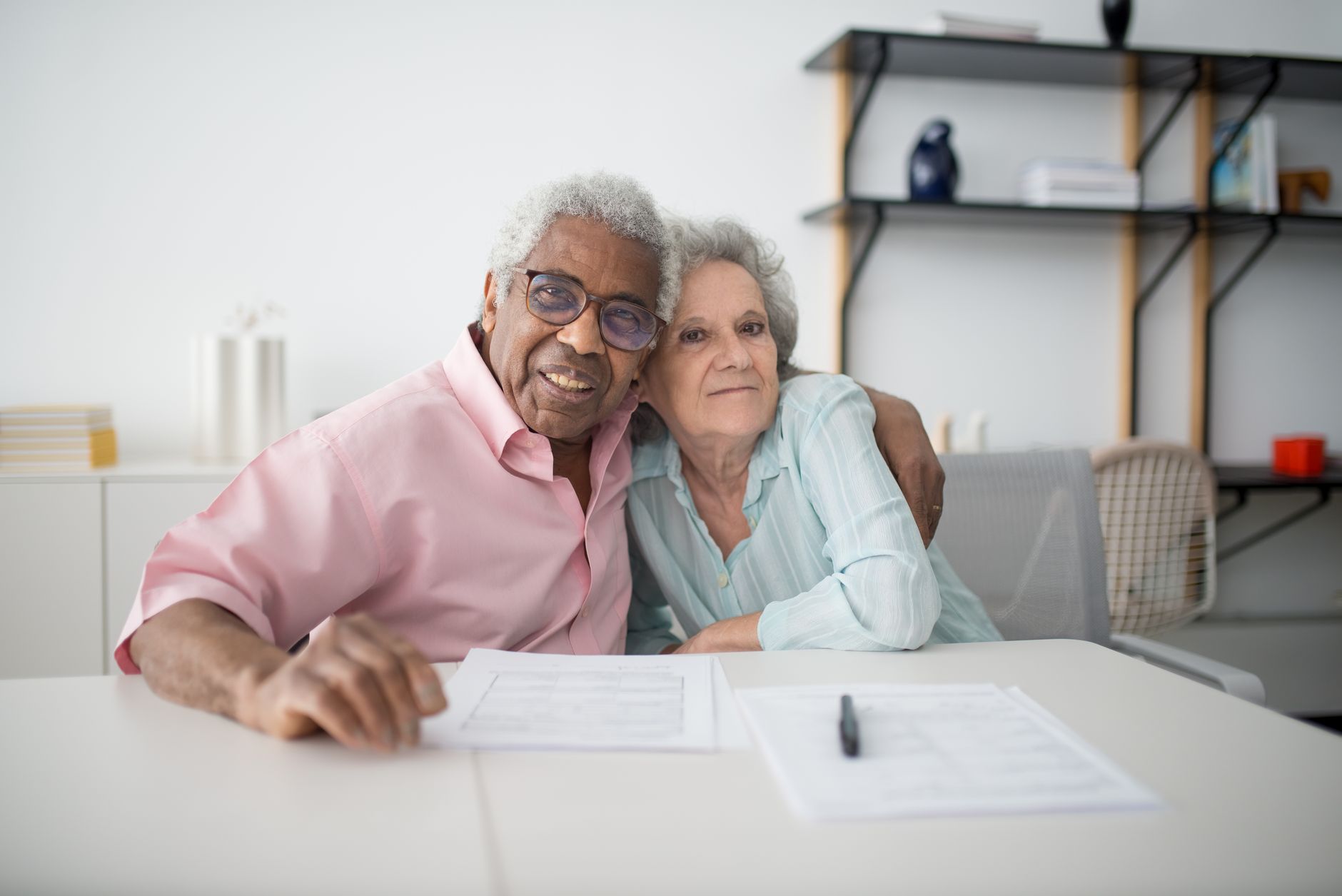 Elderly couple smiling and hugging while looking at documents on table indoors.